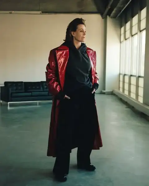 Sigourney Weaver posing in a long, vibrant red patent leather trench coat over a black hoodie and trousers in an industrial studio setting.