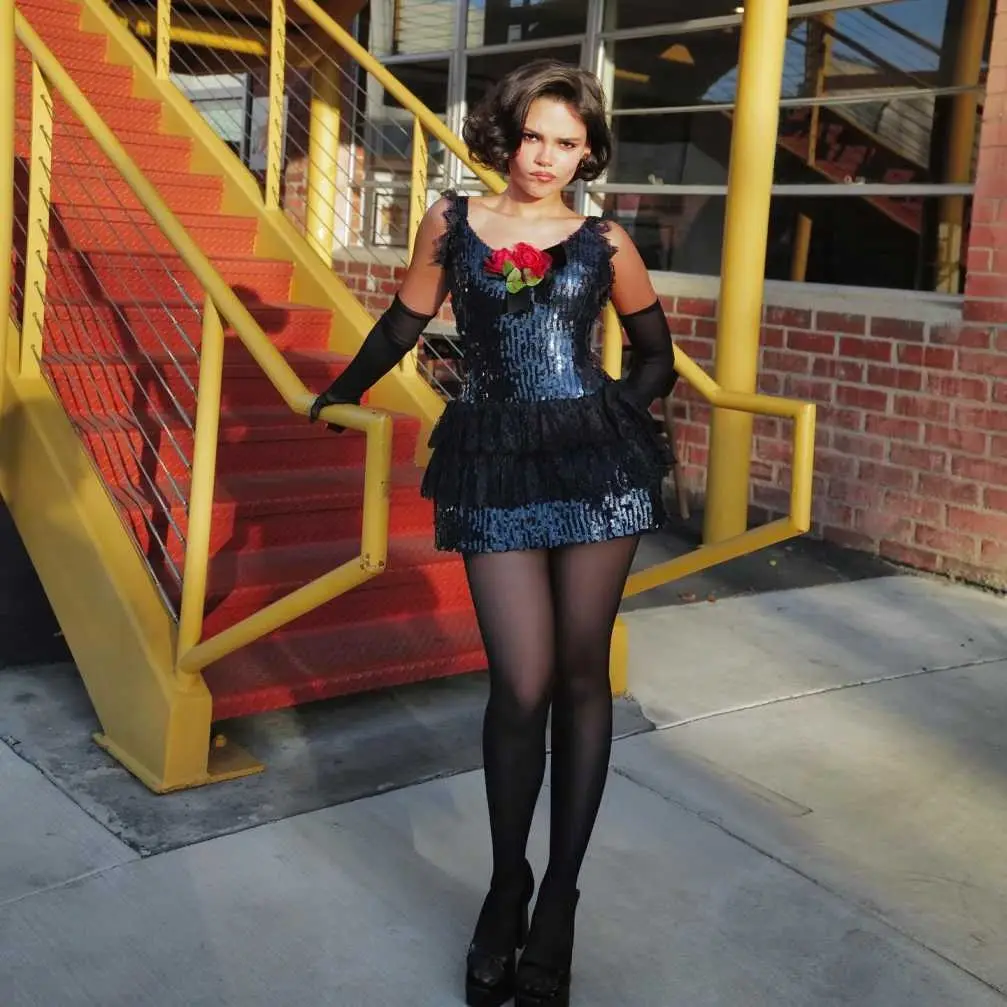 Ariana Greenblatt posing in a black sequined dress with a red rose accent beside a yellow staircase.