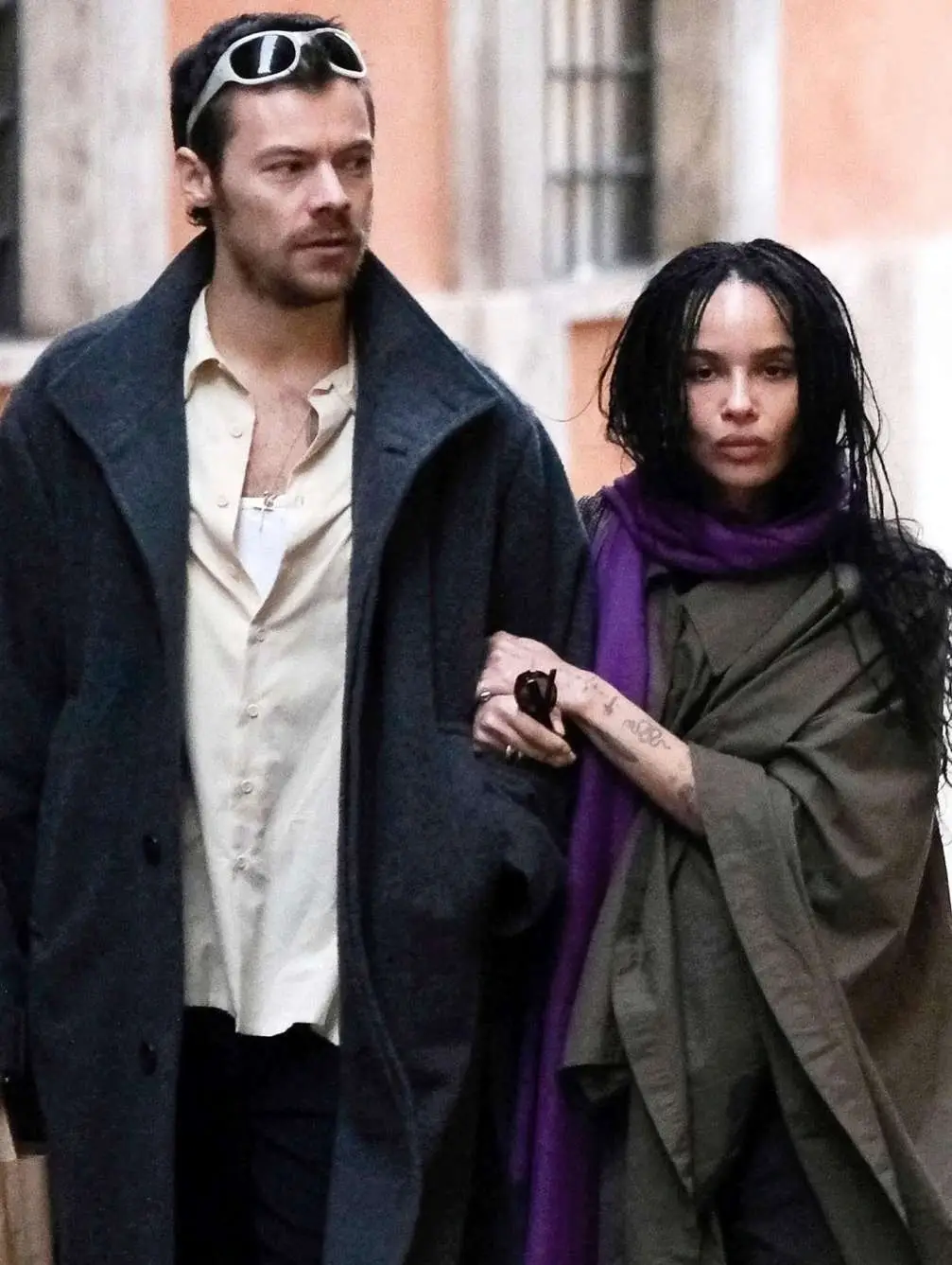 Harry Styles in a dark overcoat walking arm-in-arm with Zoë Kravitz wearing an olive poncho and purple scarf.