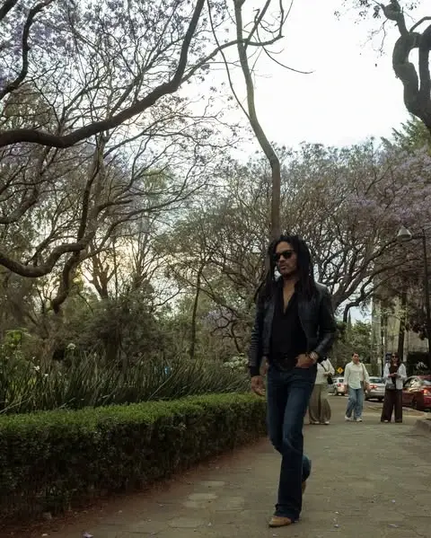 Lenny Kravitz walks along a tree-lined sidewalk wearing a black leather jacket, sunglasses, and denim jeans.