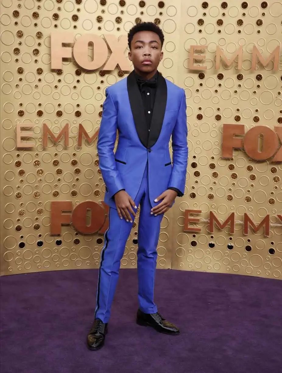 Asante Blackk posing on a purple carpet in a vibrant blue tuxedo with black lapels at the Emmy Awards.