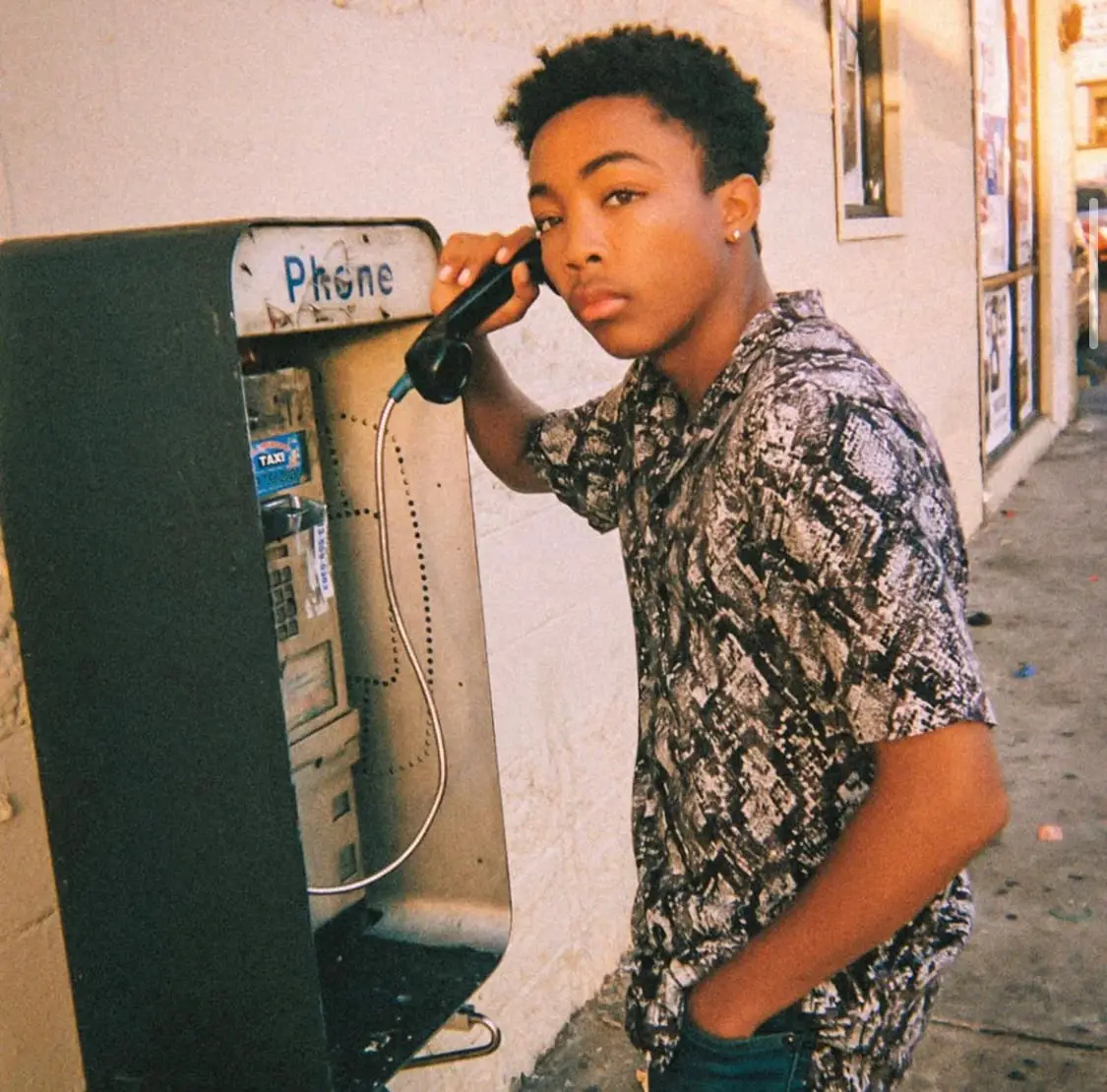 Asante Blackk posing in a patterned short-sleeve shirt while holding a vintage public payphone receiver.