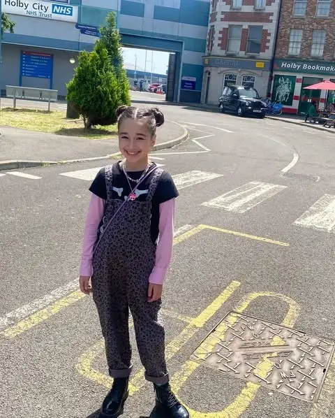 A smiling Eve Ridley wearing leopard print overalls and pink sleeves standing outdoors on a paved street.
