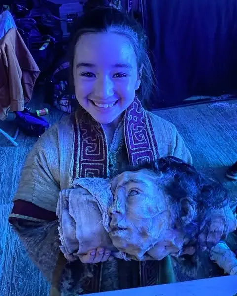 Eve Ridley smiling while holding a realistic prop head under blue lighting on a movie set.