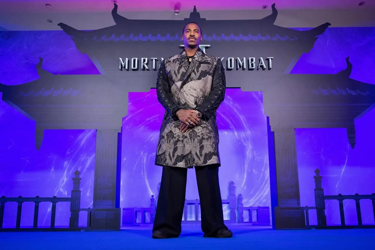 Mehcad Brooks poses in a long, patterned brocade coat in front of a Mortal Kombat themed backdrop and gate.