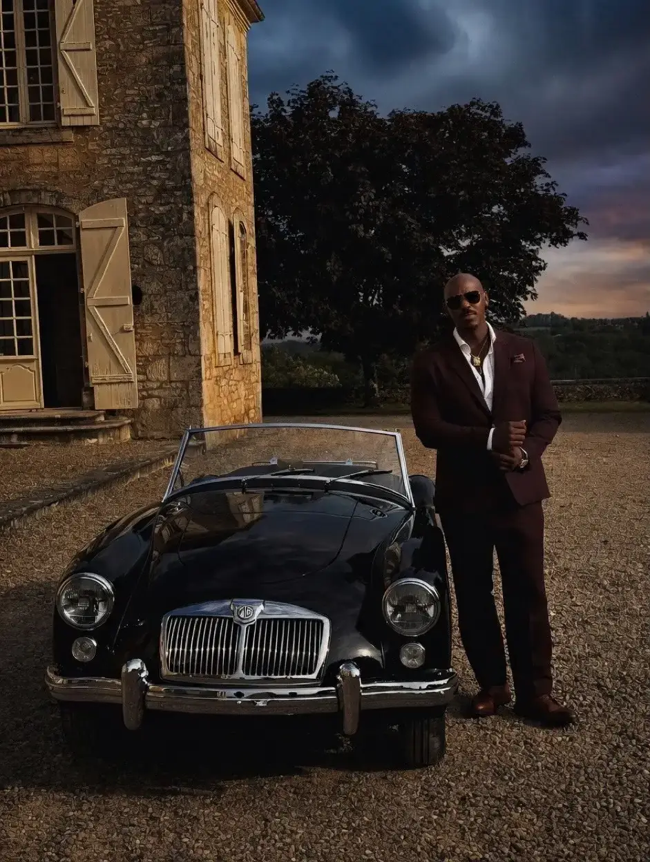 Mehcad Brooks stands in a sharp burgundy suit next to a classic black MG convertible in front of a rustic stone villa.