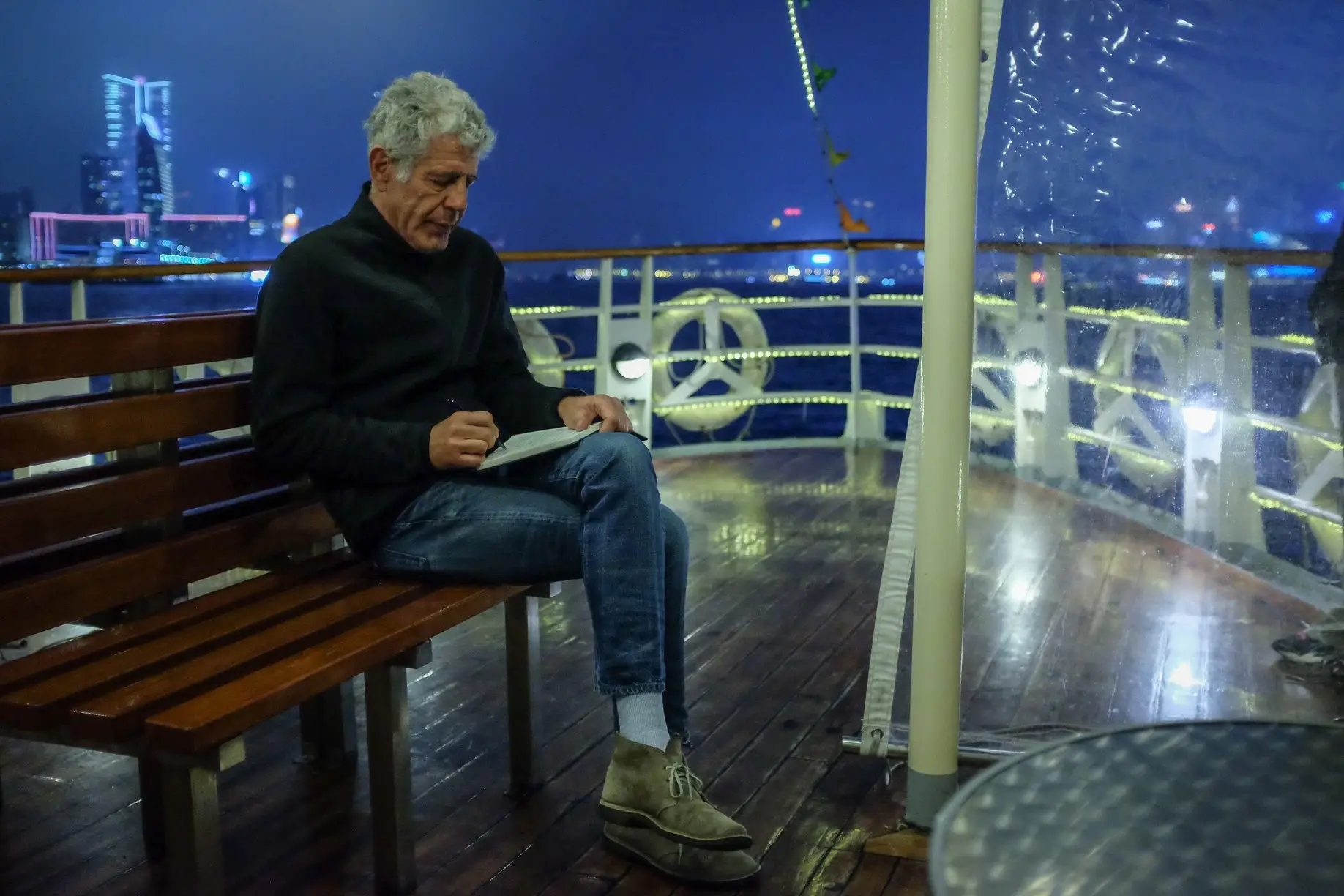 Anthony Bourdain sits on a wooden bench on a boat at night, focused on writing in a notebook with a glowing city skyline in the background.