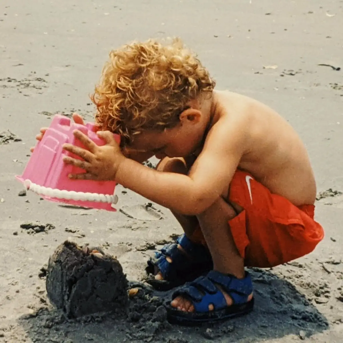 A young Michael Johnston with curly blonde hair plays with a pink sand bucket on a beach.