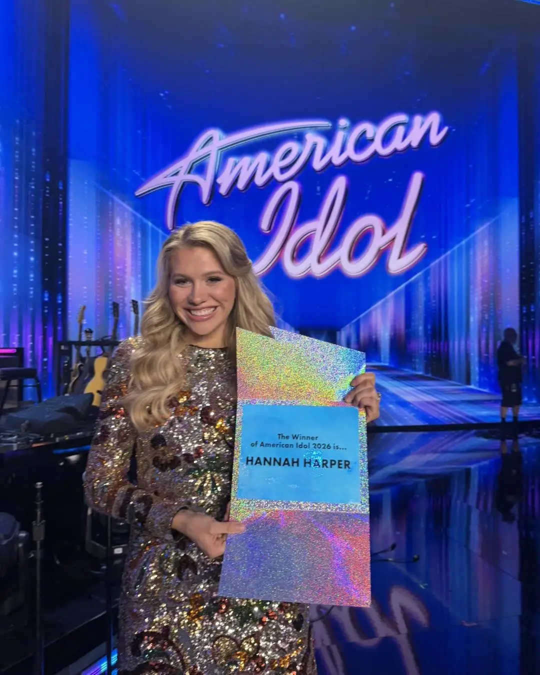 A smiling Hannah Harper in a sparkling gold dress holds a winner