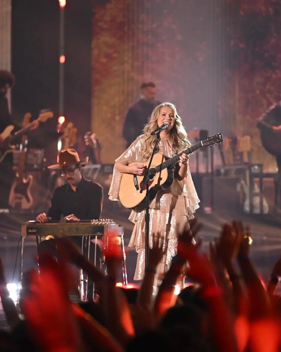 Hannah Harper performs on a brightly lit stage while playing an acoustic guitar for a cheering crowd.