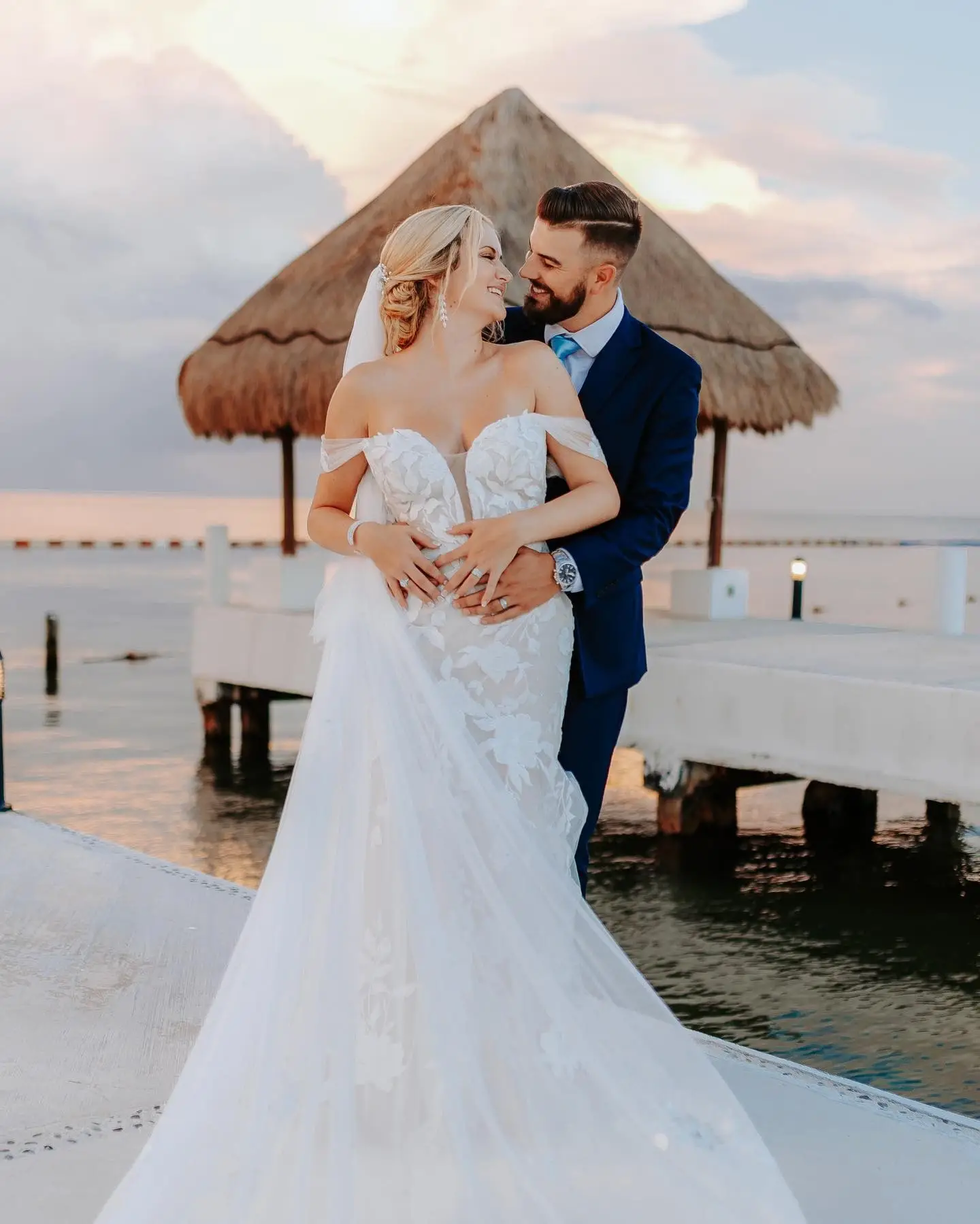 A bride and groom smile at each other on a pier by the water during sunset.
