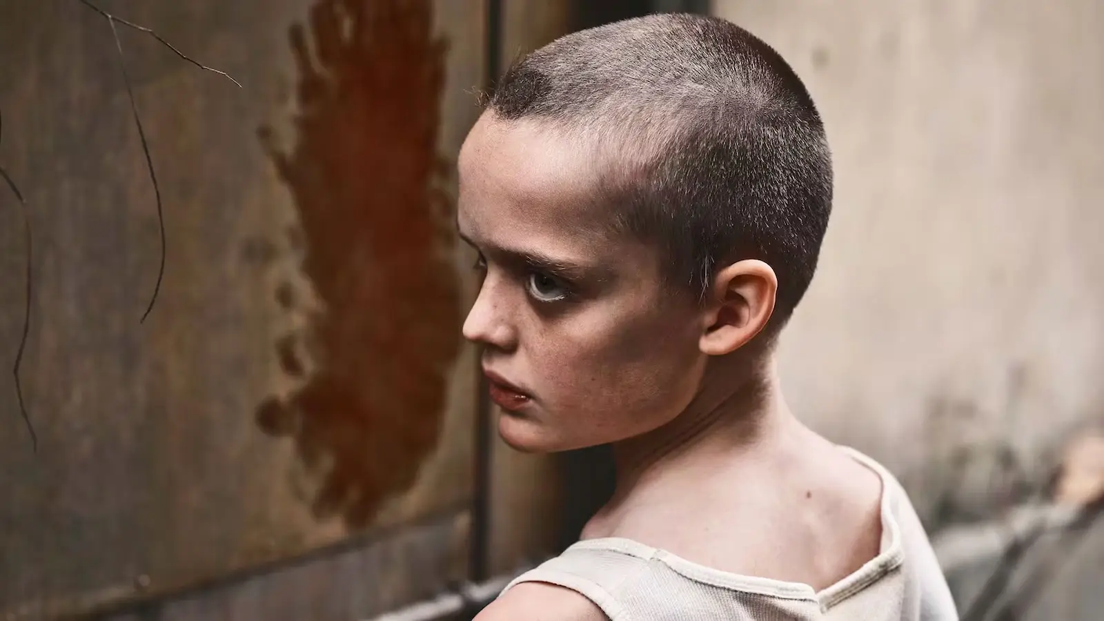 Profile view of a young boy with a shaved head and an intense expression, looking over his shoulder against a gritty, textured wall.
