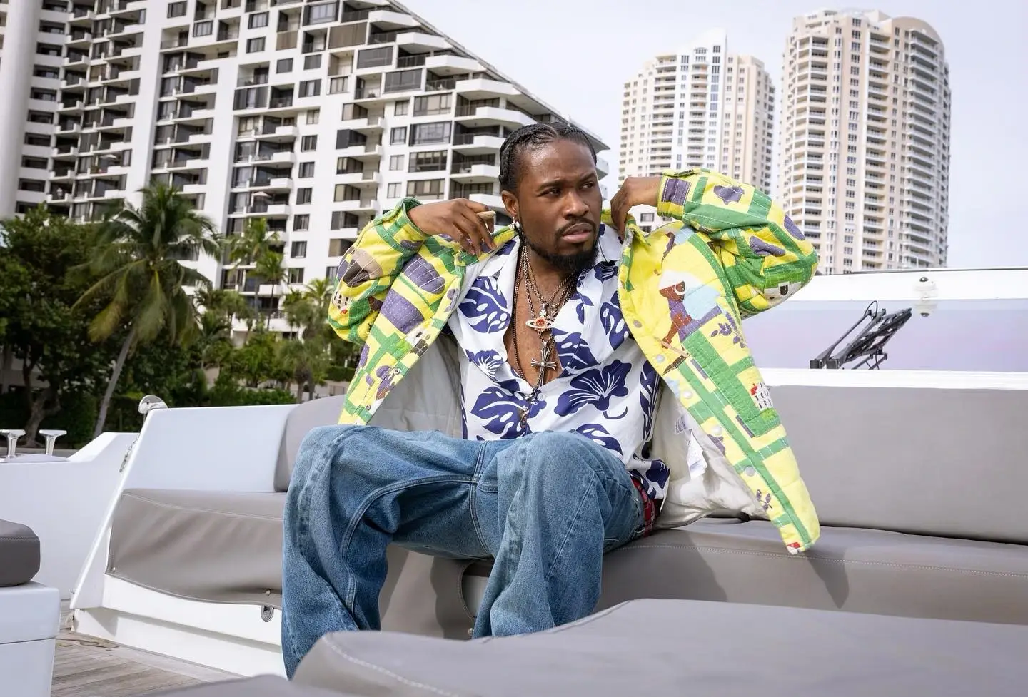 Shameik Moore sits on a yacht wearing a patterned jacket, floral shirt, and baggy jeans with high-rise buildings in the background.