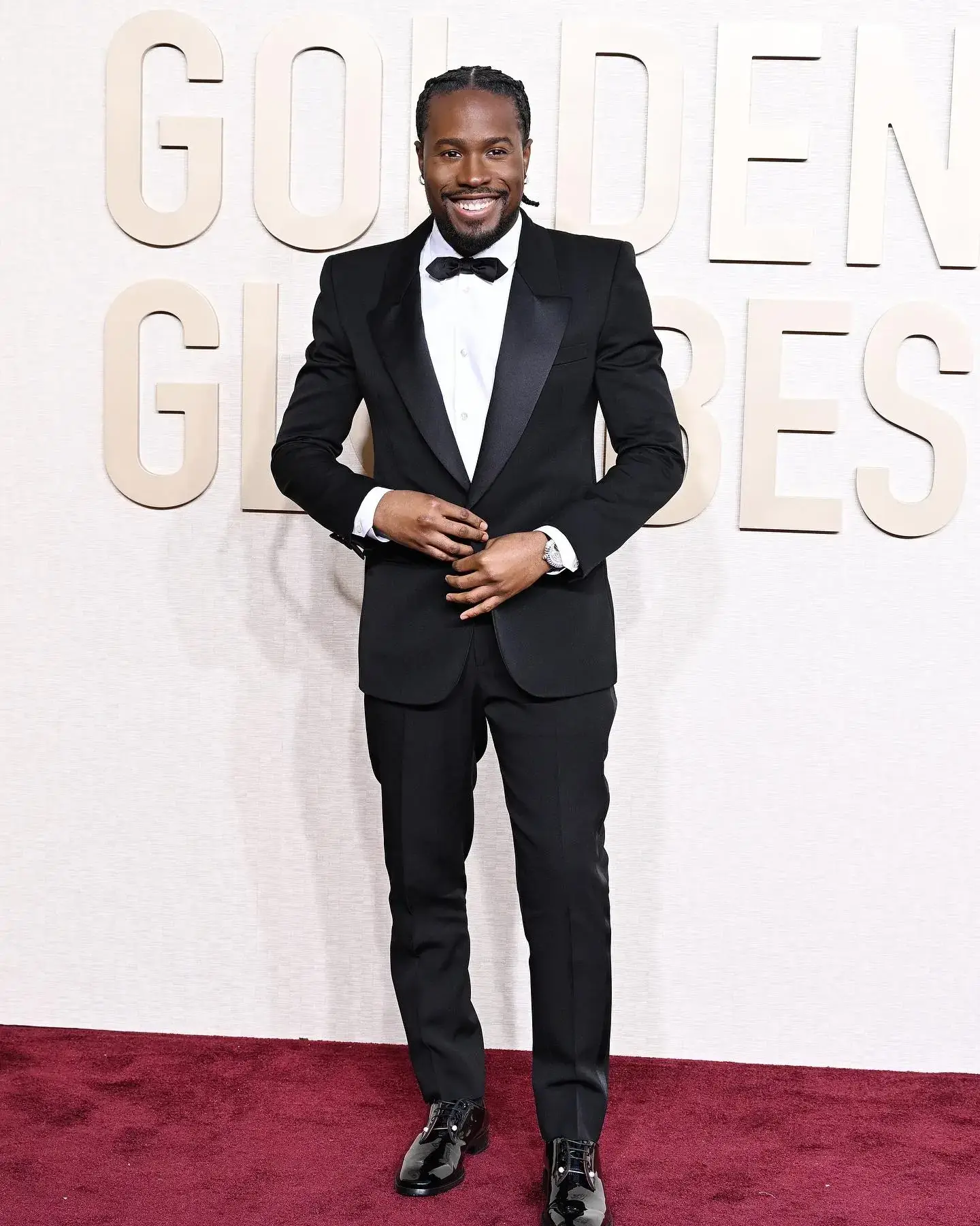Shameik Moore smiles on the red carpet in a classic black tuxedo and bow tie at the Golden Globes.