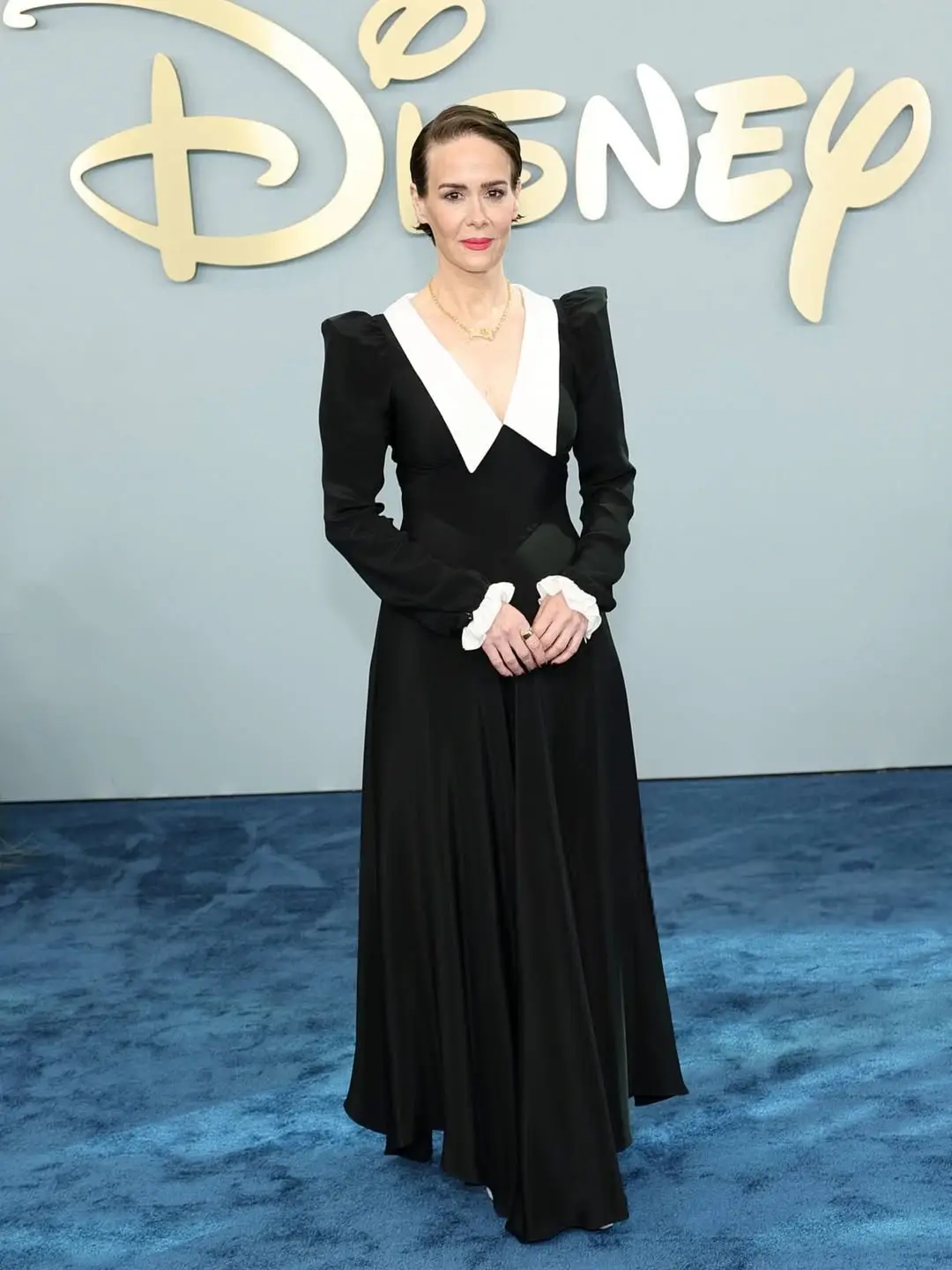 Sarah Paulson stands on a blue carpet in front of a Disney logo, wearing a long-sleeved black gown with a sharp white collar and ruffled cuffs.