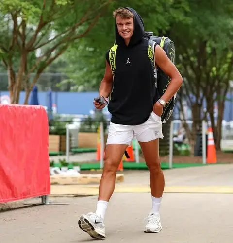 Holger Rune walking outdoors wearing a black sleeveless hoodie, white shorts, sneakers, and carrying a Babolat tennis bag.