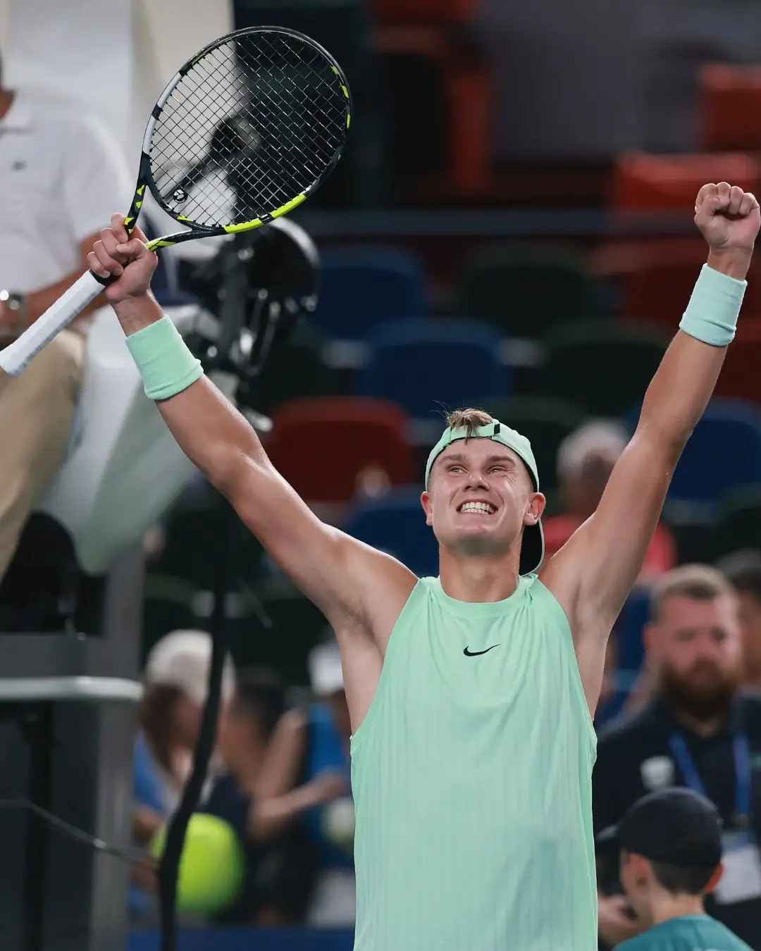Holger Rune celebrates a victory on the court with his arms raised, holding his racket in a mint green outfit.