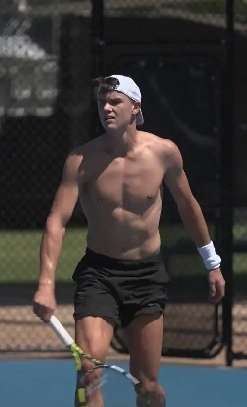 Holger Rune practices on an outdoor court wearing black shorts, a white cap, and holding a racket.