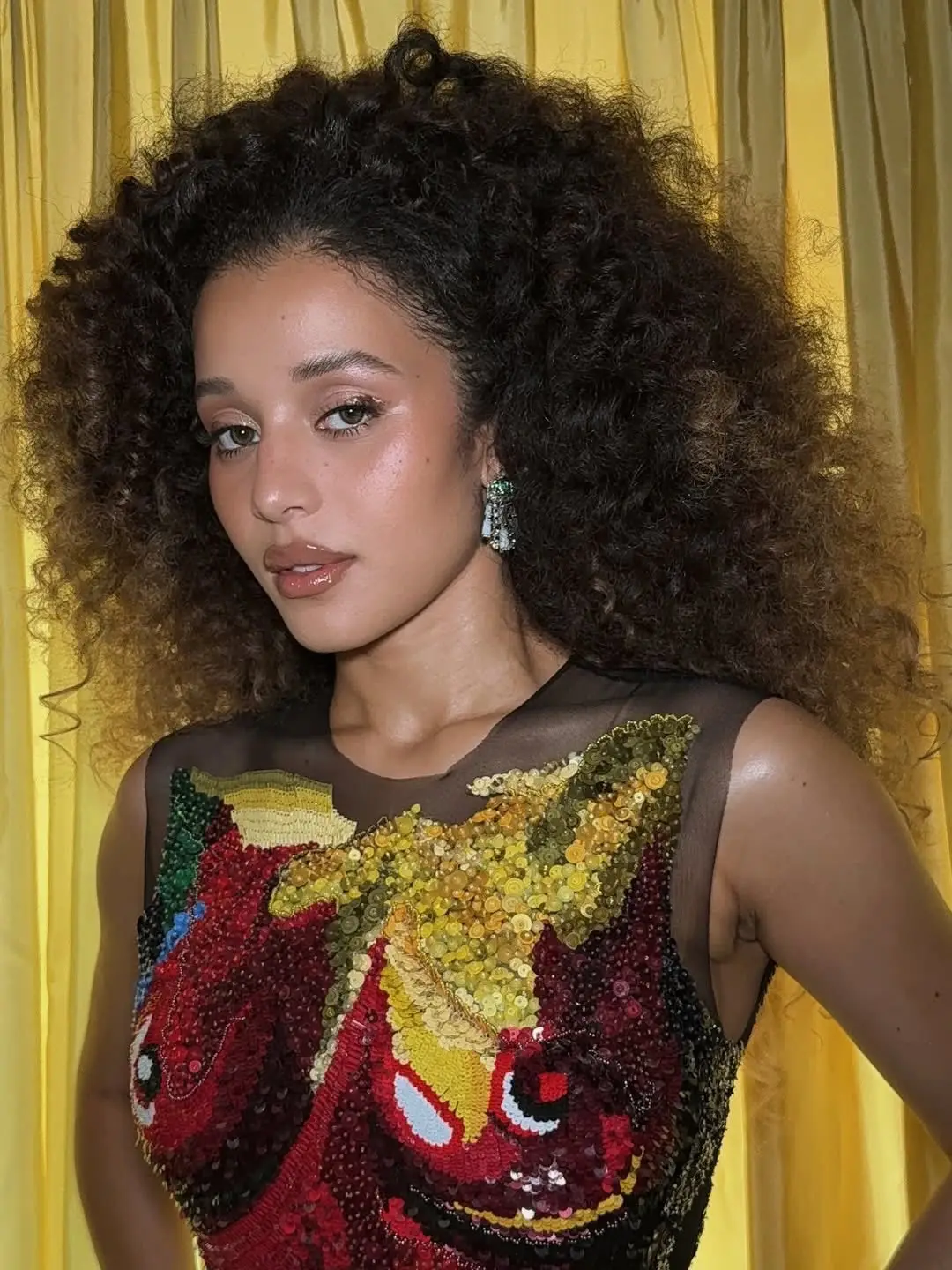 A woman with voluminous, curly brown hair and glowing makeup poses in a sheer
