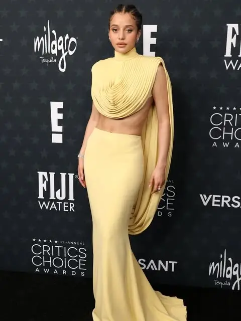 Chase Infiniti poses on a red carpet wearing a pale yellow two-piece gown featuring a pleated, draped crop top and a matching long skirt.
