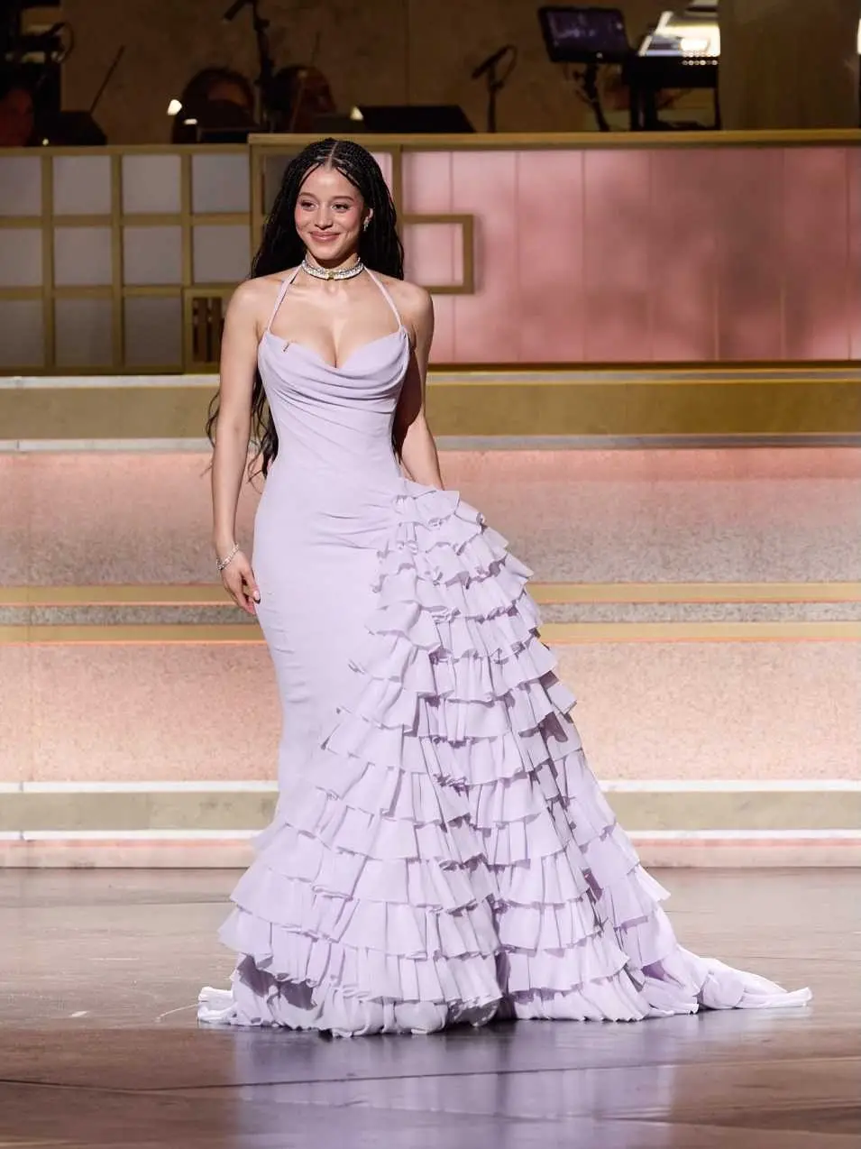 Chase Infiniti smiles on stage wearing a lavender halter gown with a dramatic tiered ruffle skirt and a choker necklace.