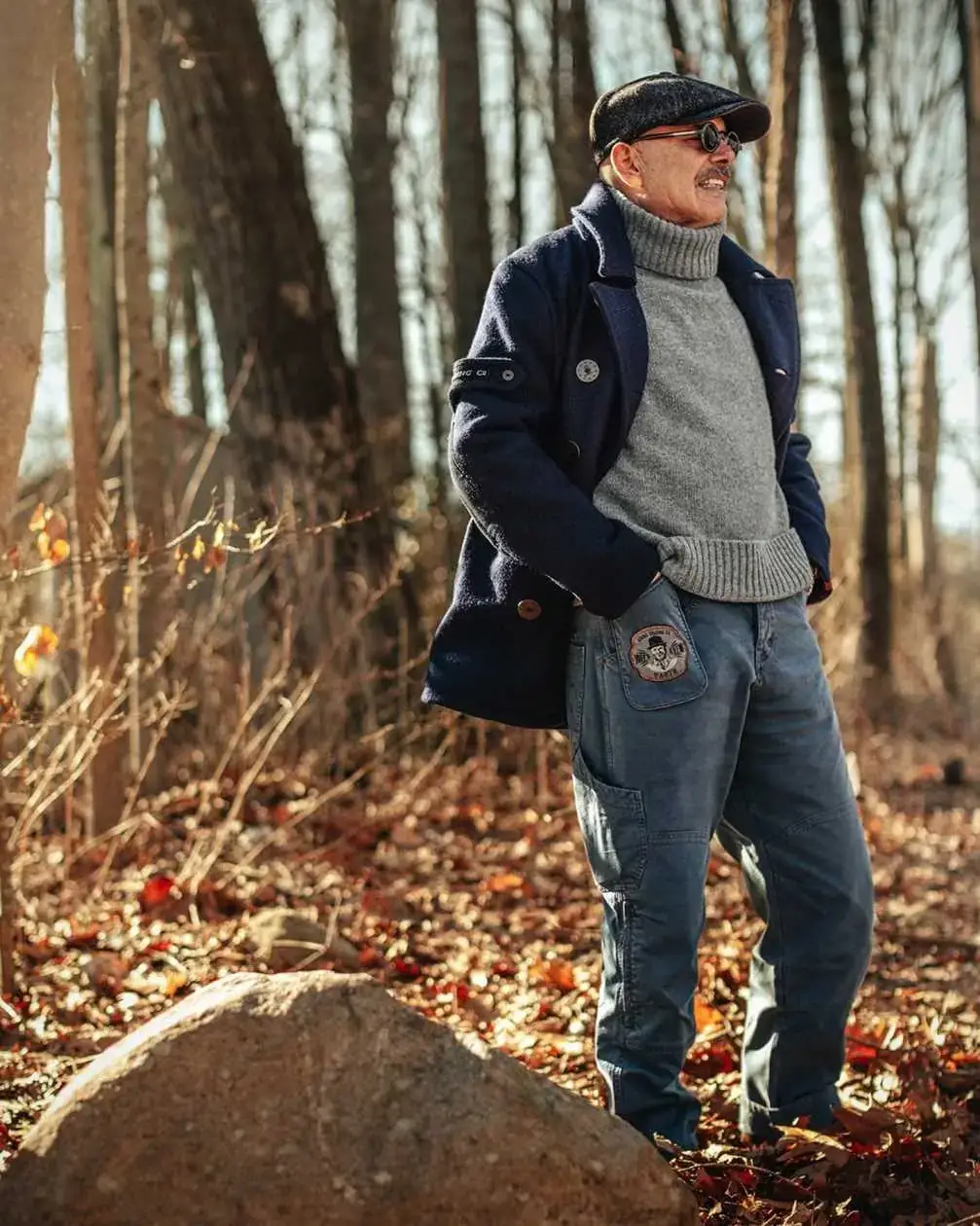 Joe Pantoliano poses in a wooded forest setting wearing a navy pea coat, grey turtleneck sweater, flat cap, and blue cargo pants.