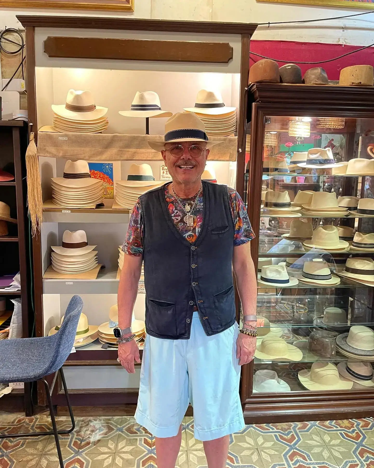 Joe Pantoliano smiles while wearing a tan fedora, dark vest, colorful shirt, and light blue shorts inside a hat boutique.