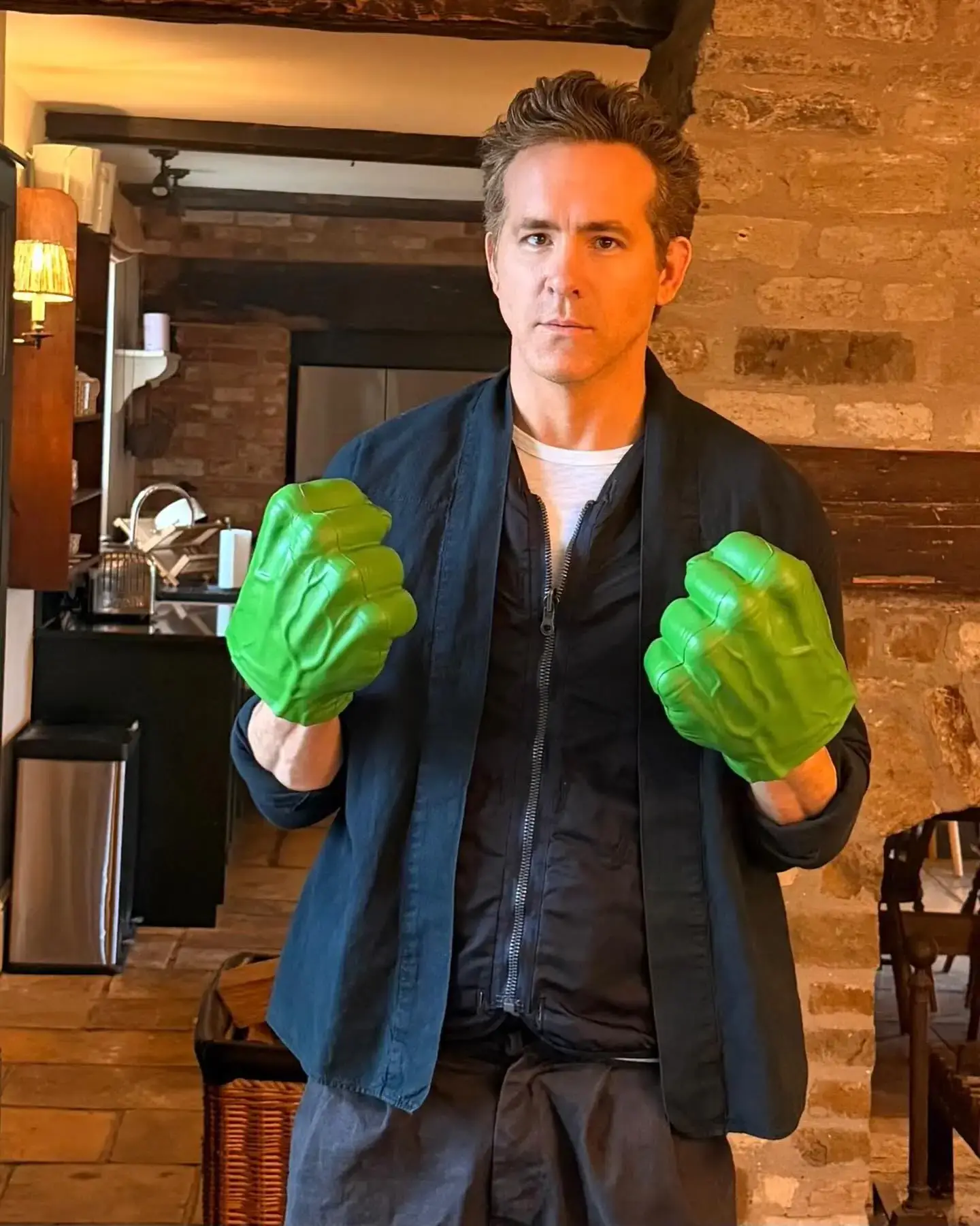 Ryan Reynolds standing in a rustic kitchen while wearing large, green Hulk-fisted gloves and posing with a serious expression.
