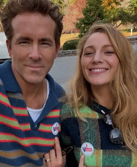 A smiling selfie of Ryan Reynolds and Blake Lively outside, both wearing sweaters and pointing to their "I Voted" stickers.