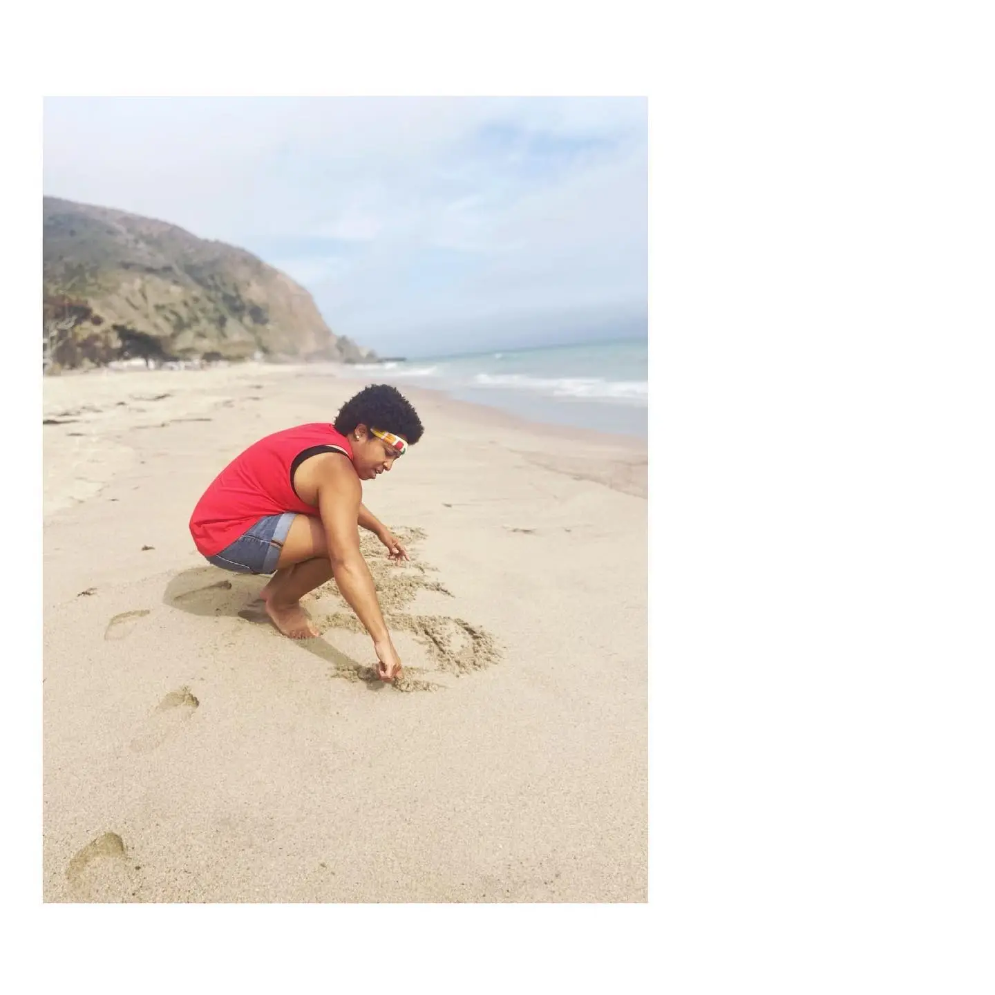 A person wearing a red tank top and denim shorts crouches on a sandy beach to write in the sand near the ocean.