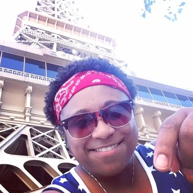 A close-up selfie of a smiling person wearing sunglasses and a red paisley bandana, with a large tower structure looming in the background.