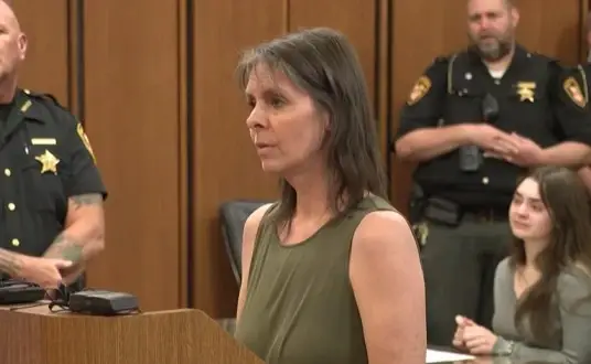 Natalie Shirilla, wearing an olive green sleeveless top, speaks at a podium in a courtroom setting with law enforcement officers standing in the background.