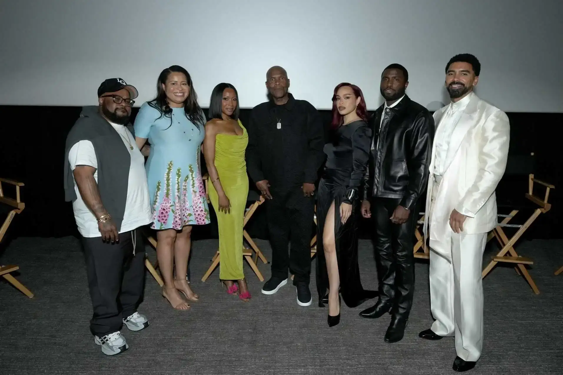The creators and cast of Netflix’s Nemesis stand together in a row on a dimly lit theater stage in front of a projection screen.