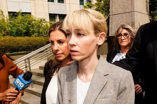 Sherri Papini walks through a crowd of reporters and microphones outside a building while wearing a grey blazer over a white shirt.