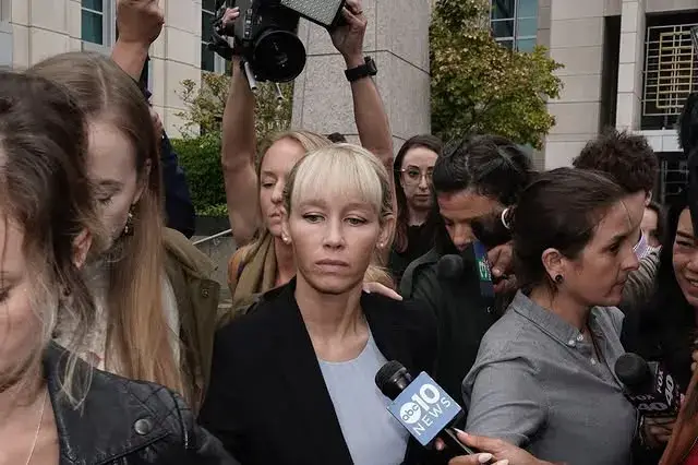 Sherri Papini walks grimly through a dense crowd of journalists holding microphones and cameras outside a building while wearing a black blazer over a light blue top.