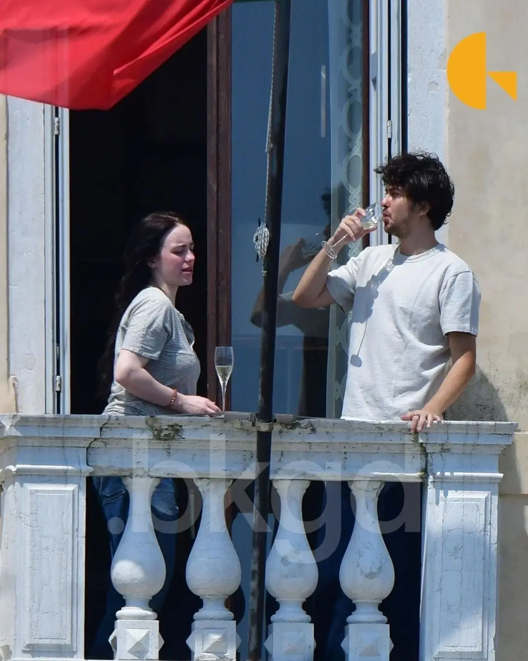 Billie Eilish and Nat Wolff stand on a stone balcony while sharing a drink and talking.