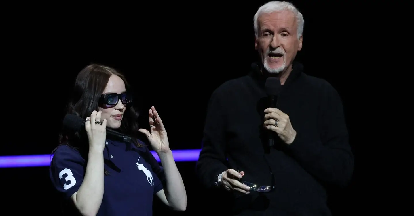 Billie Eilish adjusts her sunglasses while James Cameron speaks into a microphone next to her on stage.