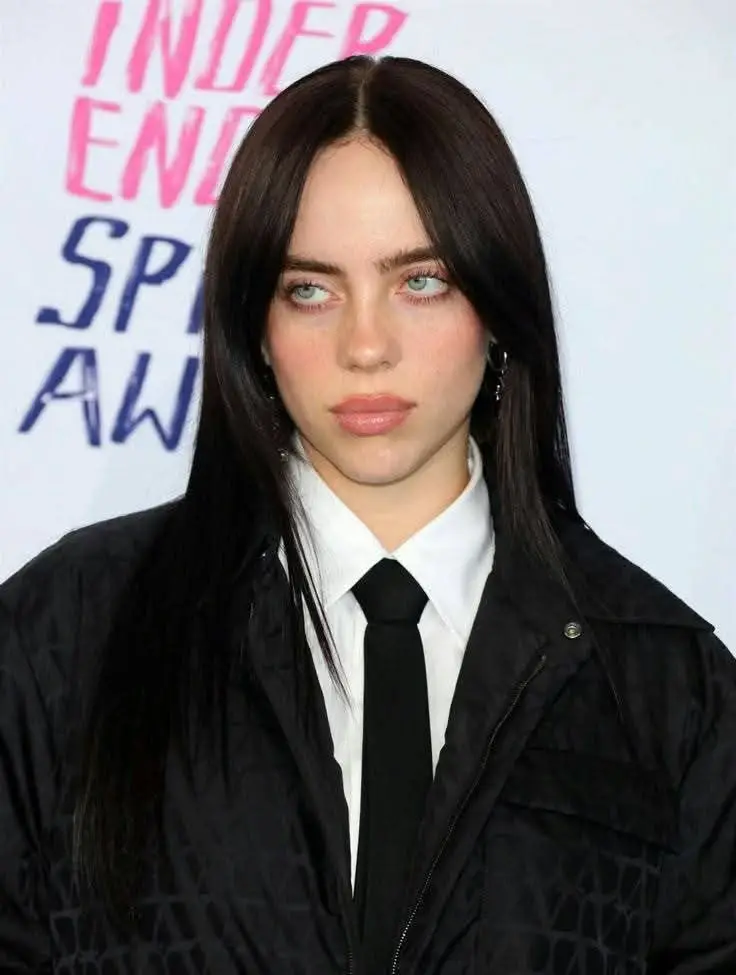 Billie Eilish poses on a red carpet wearing a black jacket, a white collared shirt, and a black tie.