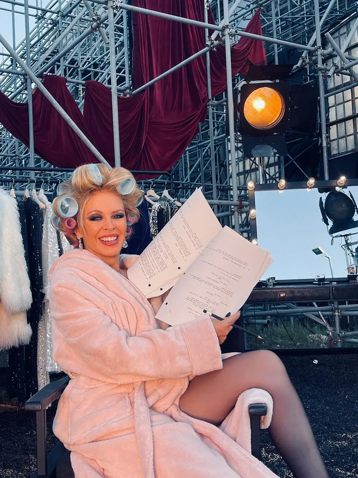 Kylie Minogue wears a pink robe and hair rollers while sitting backstage holding an open script in front of an outdoor set structure.