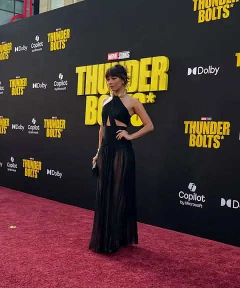 Xochitl Gomez poses on a red carpet in a halter-neck black gown with sheer paneling in front of a yellow "Thunderbolts*" backdrop.
