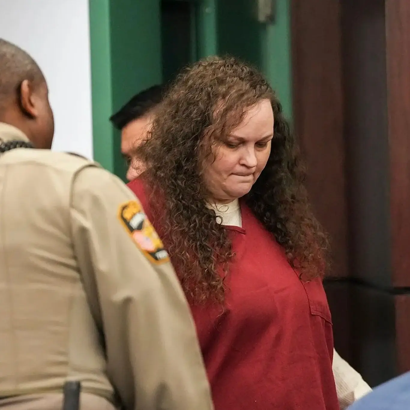 Magen Fieramusca, wearing a red jail uniform, looks downward while standing in a courtroom next to guards.