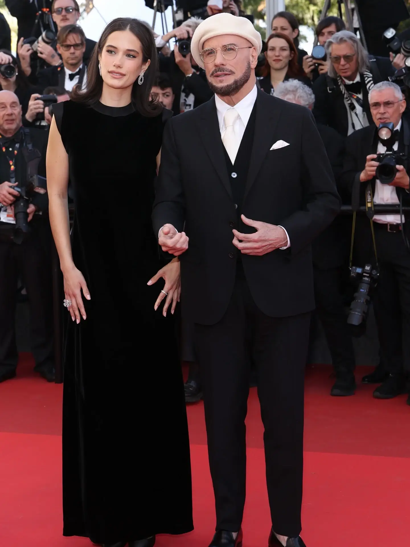 John Travolta, wearing a white beret and a black suit, stands alongside his daughter, Ella Bleu Travolta, in a black velvet gown on a red carpet.