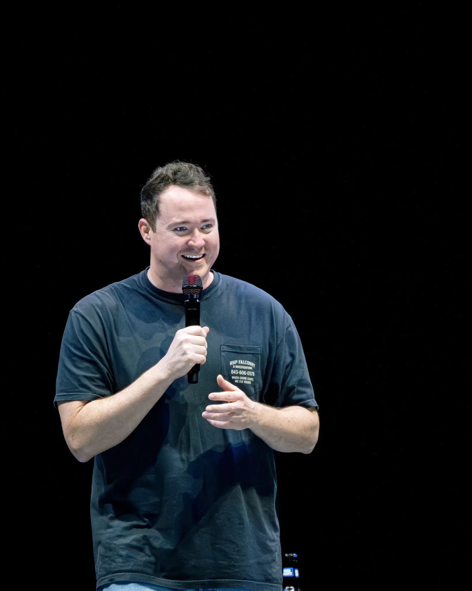 A smiling man on a dark stage holds a microphone and gestures while speaking.