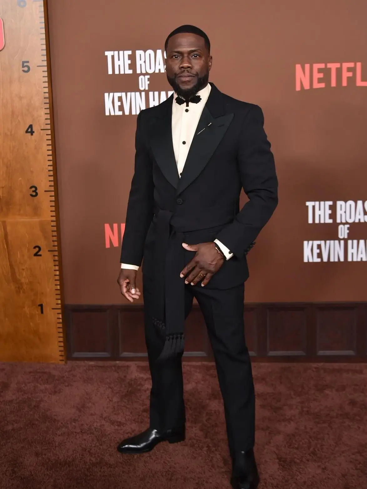 Kevin Hart poses on a brown carpet wearing a black tuxedo jacket with a tied sash and a bow tie.