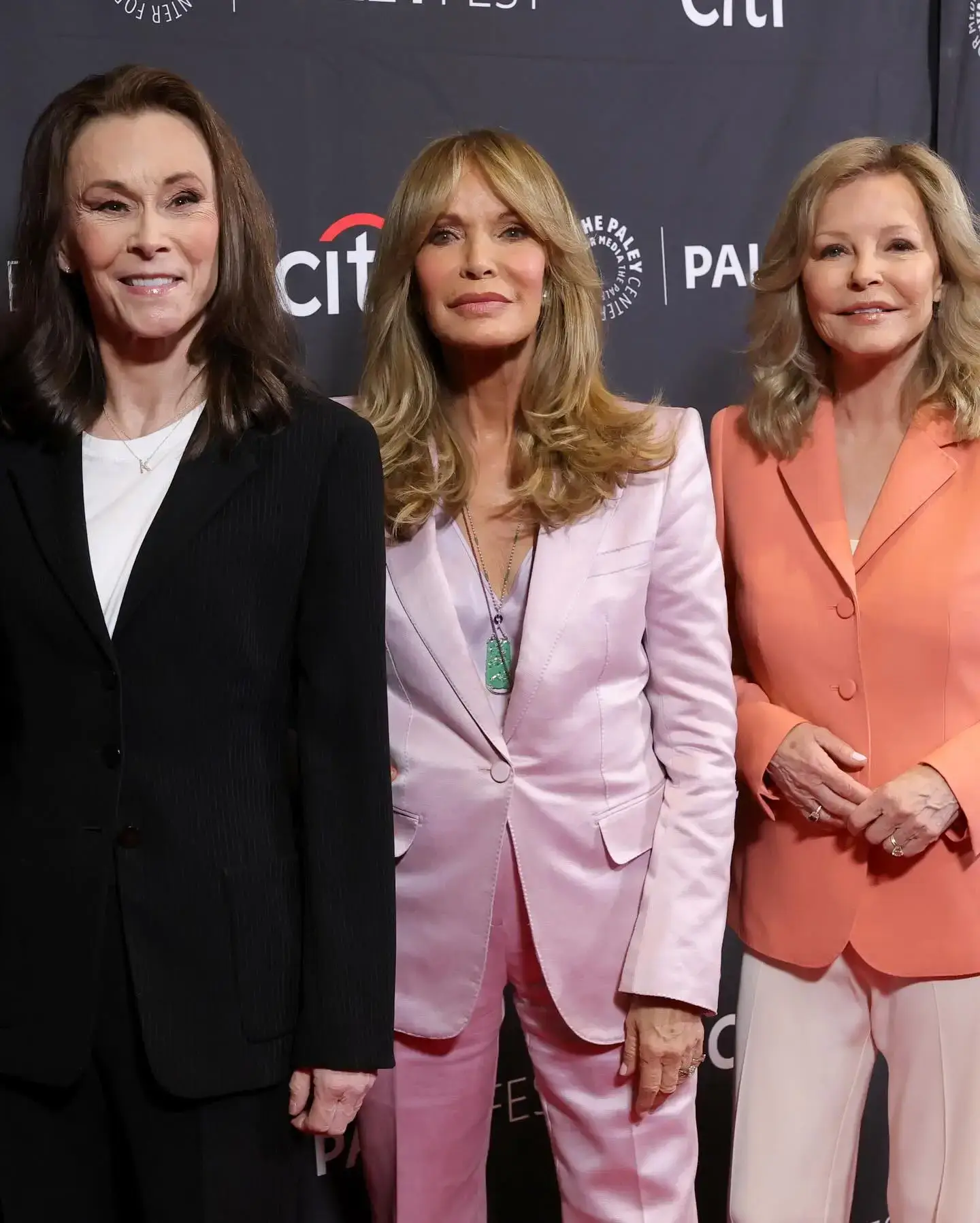 Kate Jackson in a black blazer, Jaclyn Smith in a lavender satin pantsuit, and Cheryl Ladd in a peach blazer smile together at a red carpet event.