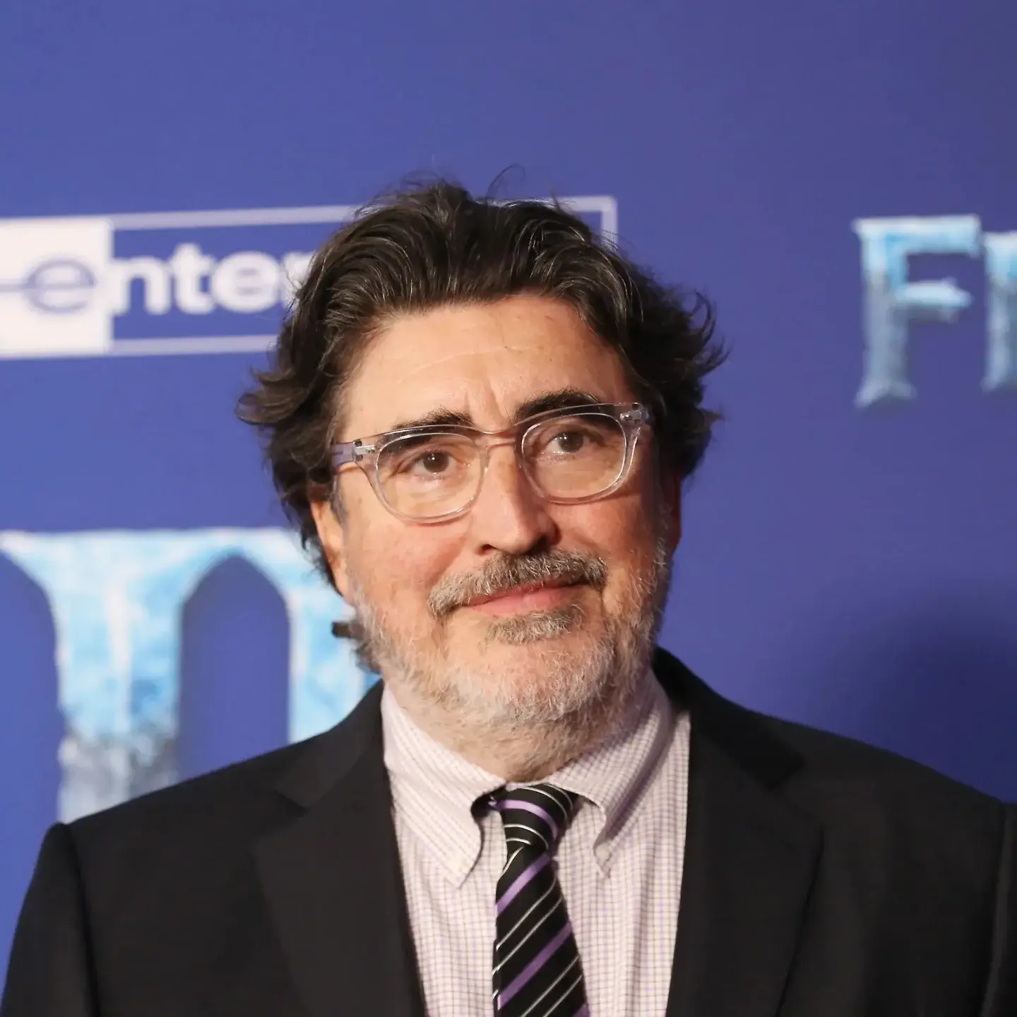 Alfred Molina smiles while wearing clear-framed glasses, a dark suit jacket, and a striped tie in front of a blue backdrop.