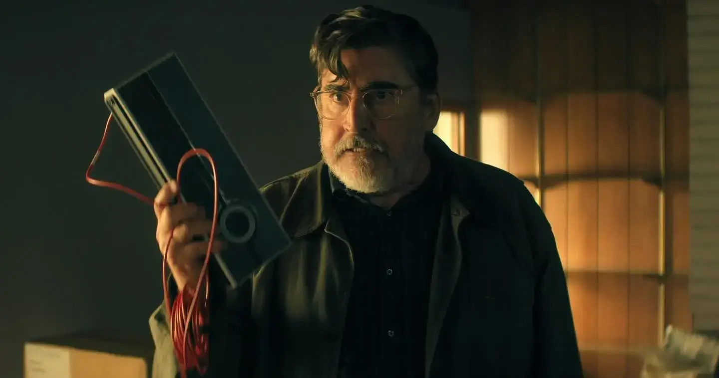 Alfred Molina looks intensely ahead while holding up a metallic device with a red cord in a dimly lit room.