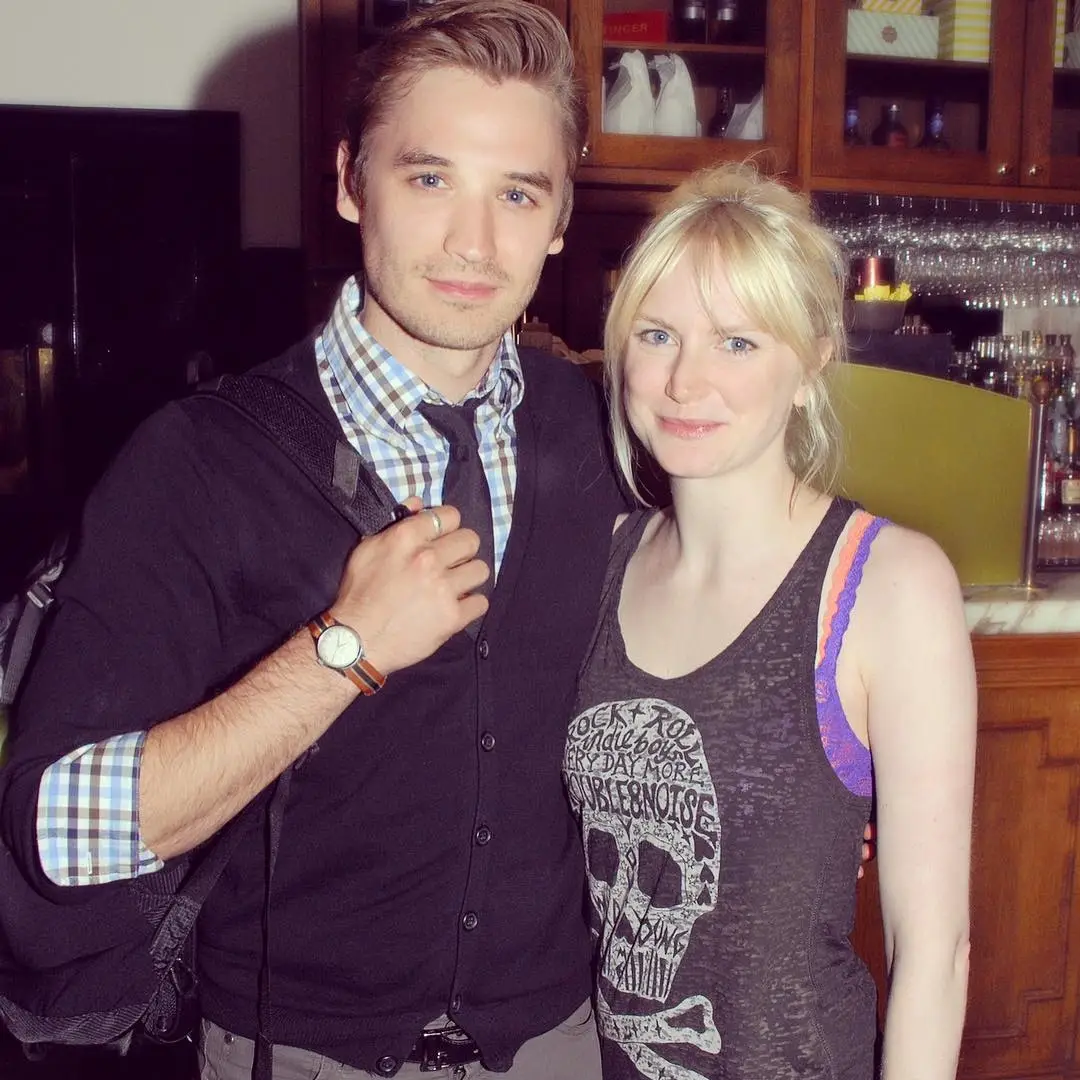 eth Numrich stands with his arm around actress Louise Dylan while posing together indoors.