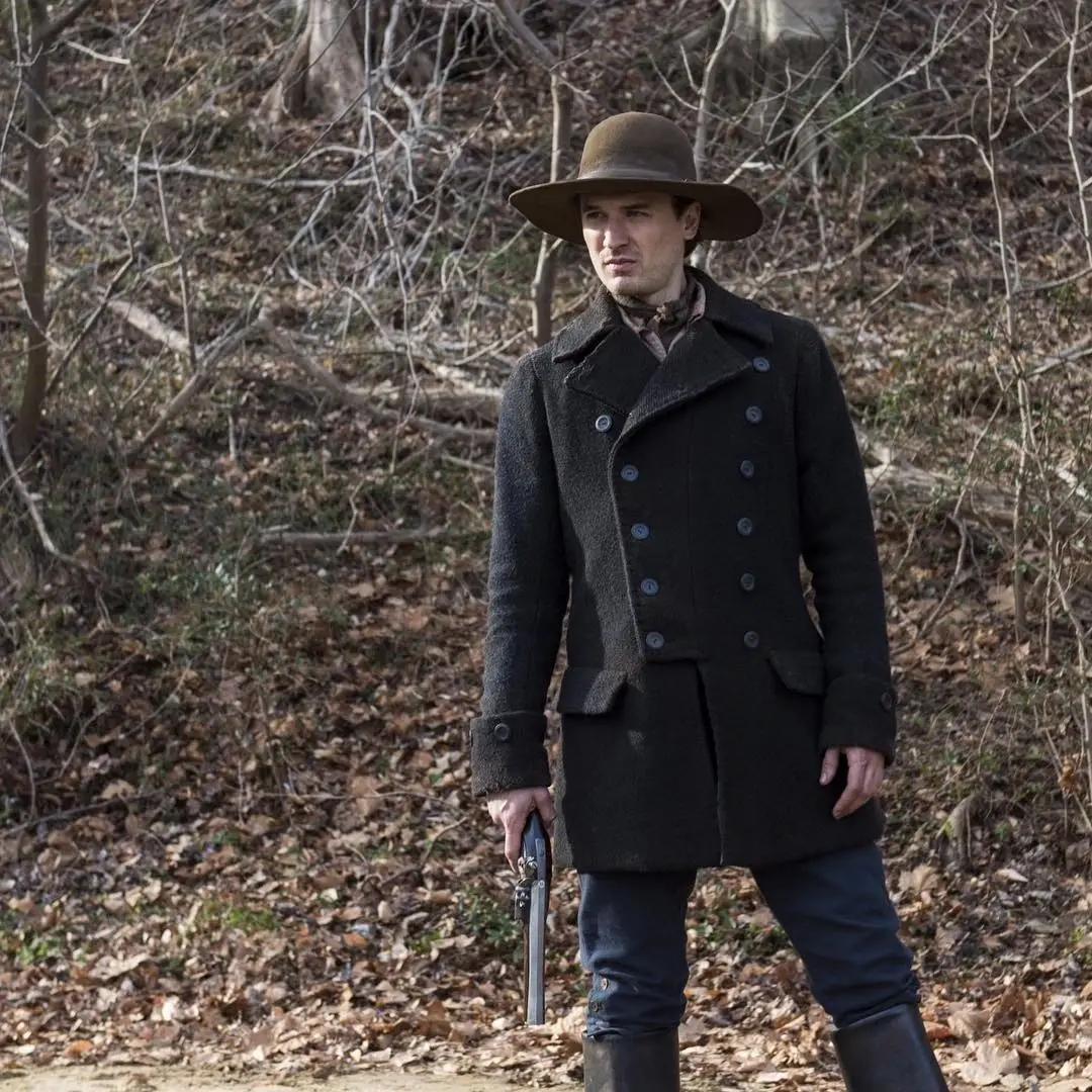 Seth Numrich stands outdoors in a wooded area wearing a historical coat and hat while holding a flintlock pistol.