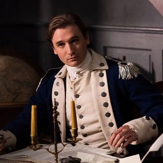 Seth Numrich sits at a desk wearing an 18th-century military uniform with epaulets next to a candle holder.